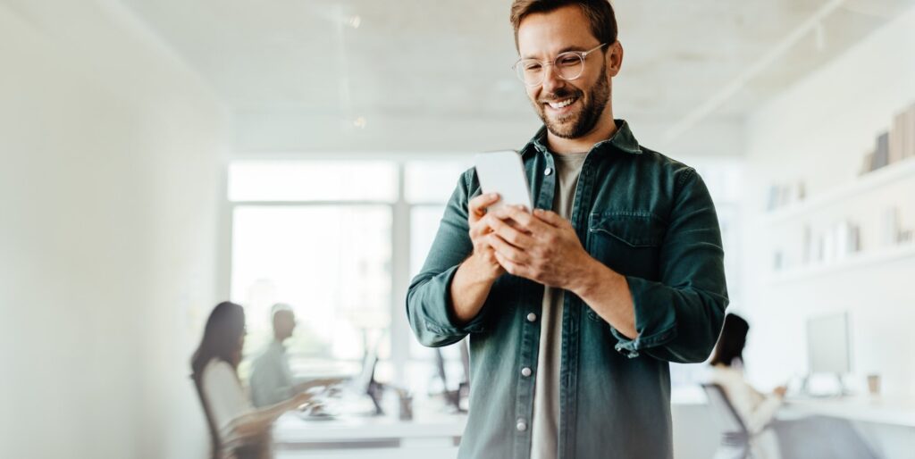 Business man reading a message on a mobile phone in an office. Male business professional standing in a co-working office.