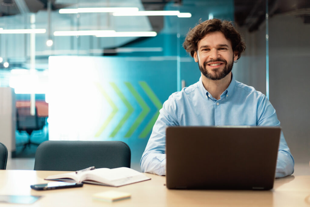 People And Technology. Portrait of cheerful smiling man sitting at desk in modern coworking office, looking and posing at camera. Happy middle aged guy using laptop and working, free copy space
