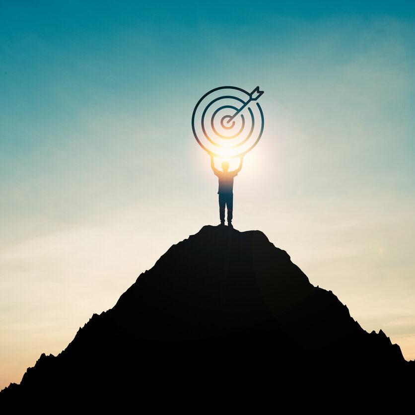 Silhouette of businessman holding target board on the top of mountain with over blue sky and sunlight. It is symbol of leadership successful achievement with goal and objective target.